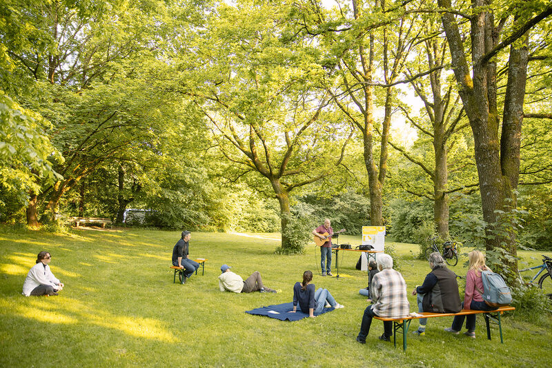 Mehrere Menschen sitzen in der Parkmeile Trudering-Neuperlach und hören einem Gitarrenspieler zu.