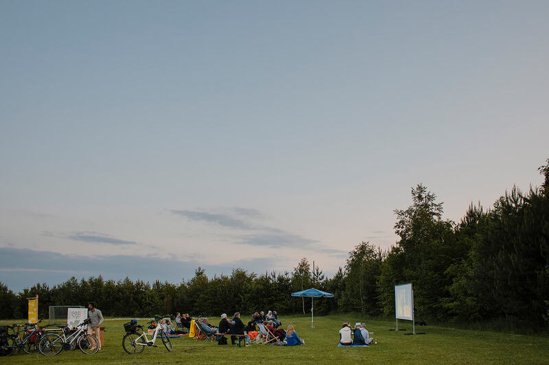 Mehrere Menschen sitzen auf Decken, Bänken und Klappstühlen vor dem Freiluftkino auf der Festwiese im Gefilde