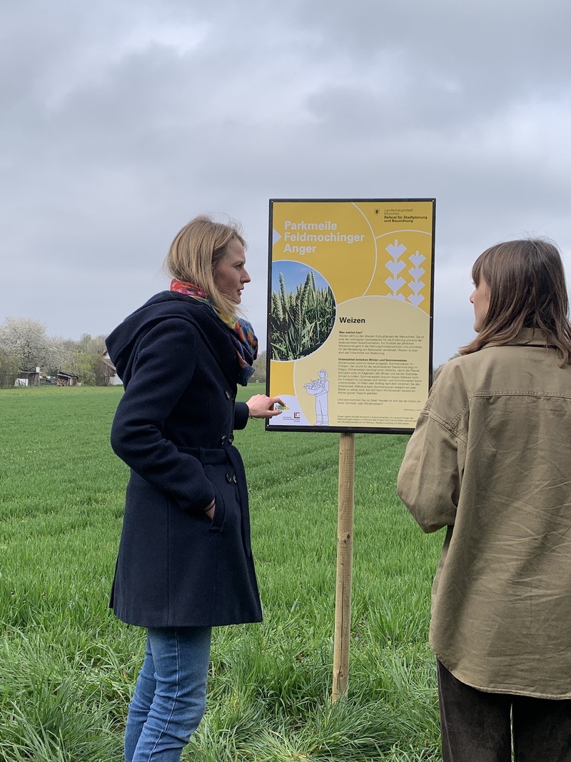 2 Personen stehen vor dem Schild des Agrarlehrpfades und reden miteinander