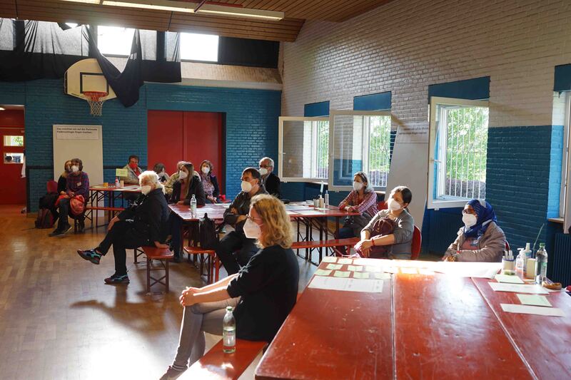 Mehrere Menschen sitzen auf Bänken und verfolgen die Eröffnung der Ideenwerkstatt in den Räumlichkeiten des Jugendtreff „s’Dülfers“.