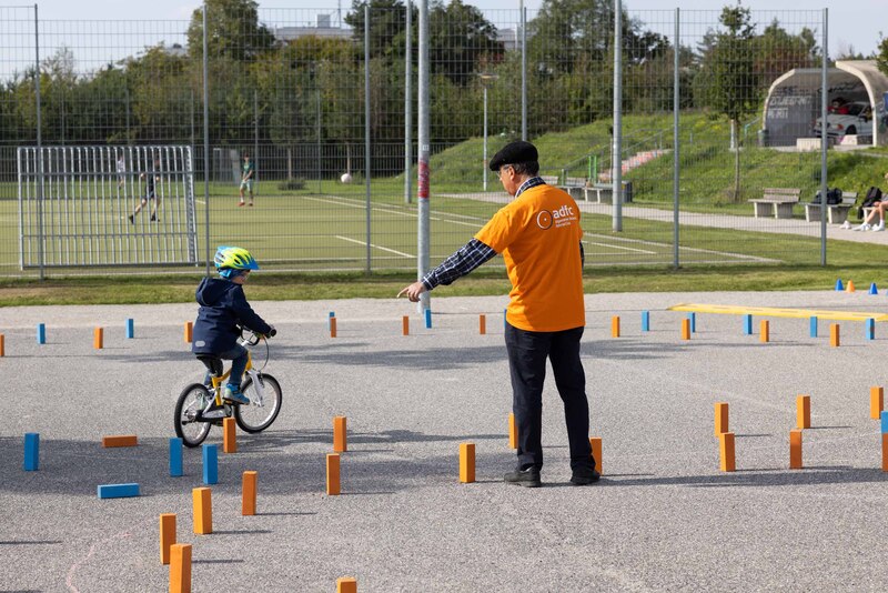 Ein Kind fährt unter Anleitung eines Ordners durch ein Fahrrad Parkour, markiert mit orangenen und blauen Holzklötzen. 